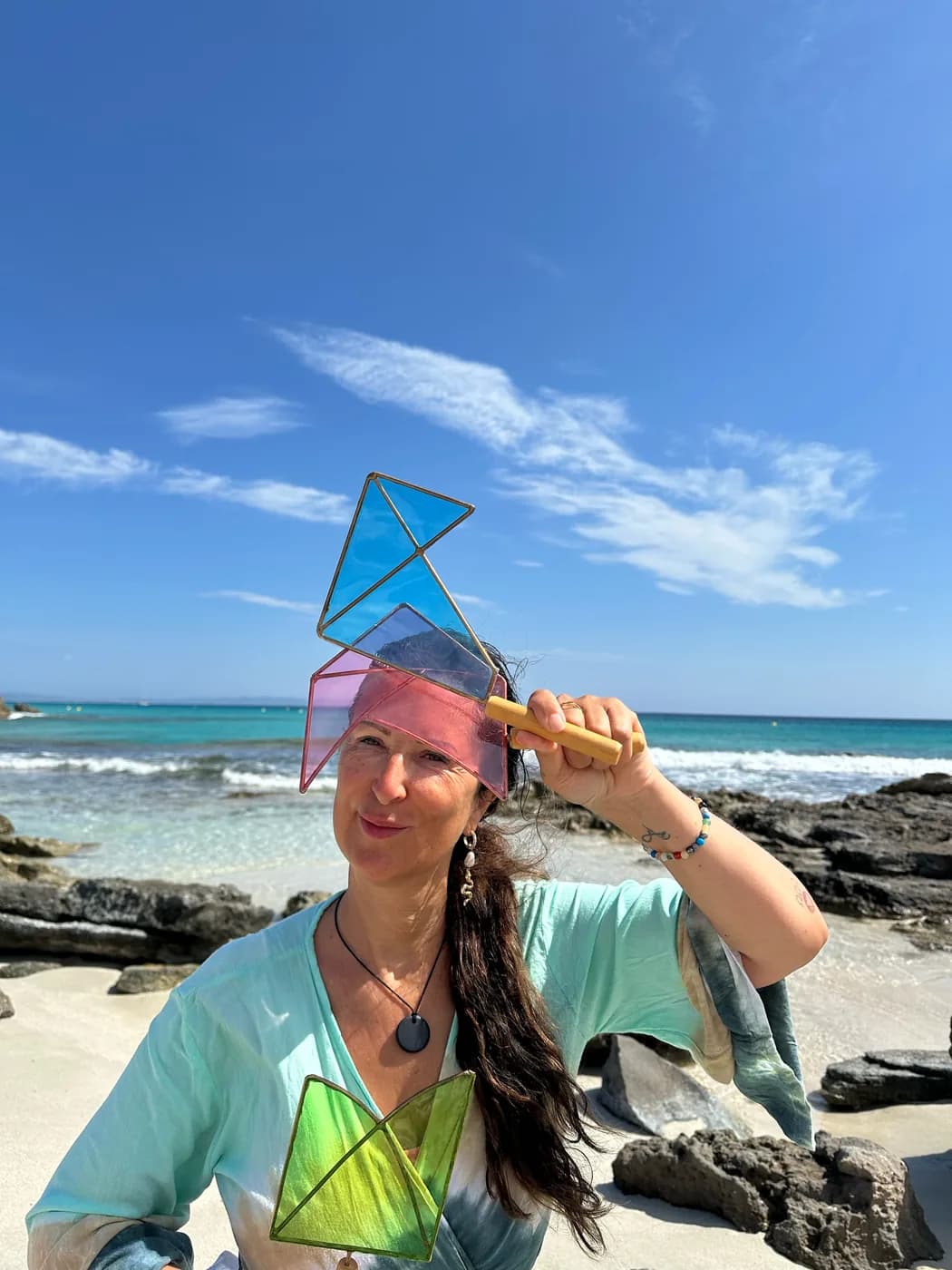 Petra Steffen an einem Strand, die ein Paddle — eine geometrische Rahmenform mit farbiger Füllung an einem Holzgriff — vor die Sonne hält.